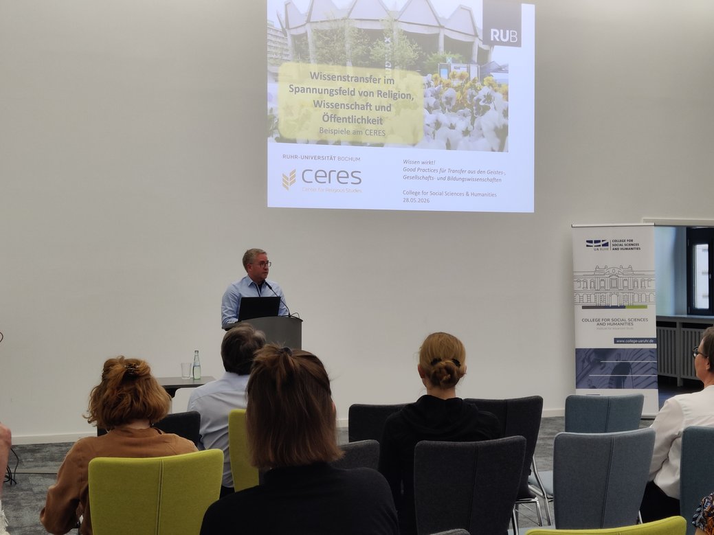 Eine Person steht an einem Rednerpult in einem modernen Hörsaal und hält einen Vortrag vor Publikum. Auf der großen Leinwand ist eine Präsentationsfolie der Ruhr-Universität Bochum bzw. des CERES – Center for Religious Studies zu sehen.